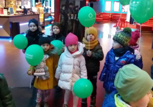 Widok na dzieci trzymające w rękach zielone baloniki. W tle widać plakaty i afisze kinowe.
